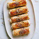 Golden, crispy phyllo feta rolls served on an oval plate, garnished with honey, nuts, and sesame seeds.