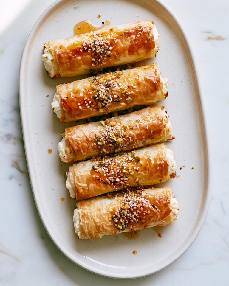 Crispy Phyllo Feta Rolls with Hot Honey & Pistachios Golden, crispy phyllo feta rolls served on an oval plate, garnished with honey, nuts, and sesame seeds.