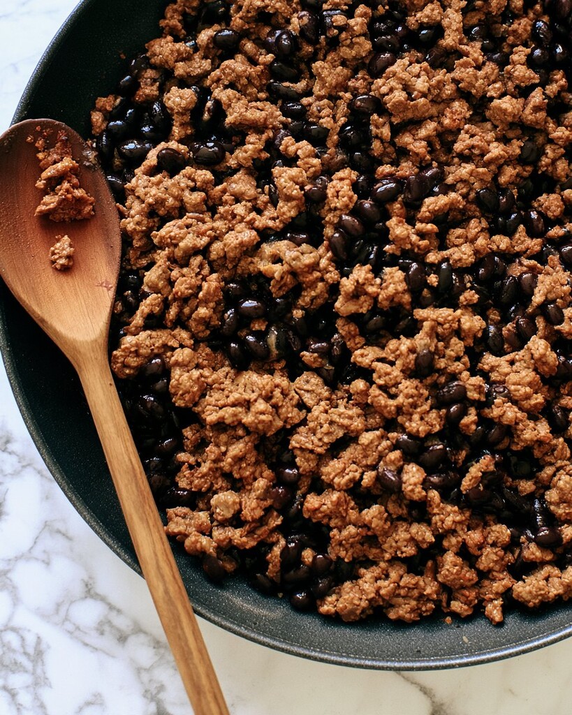 Ground beef and black beans simmering in a dark skillet with a wooden spoon