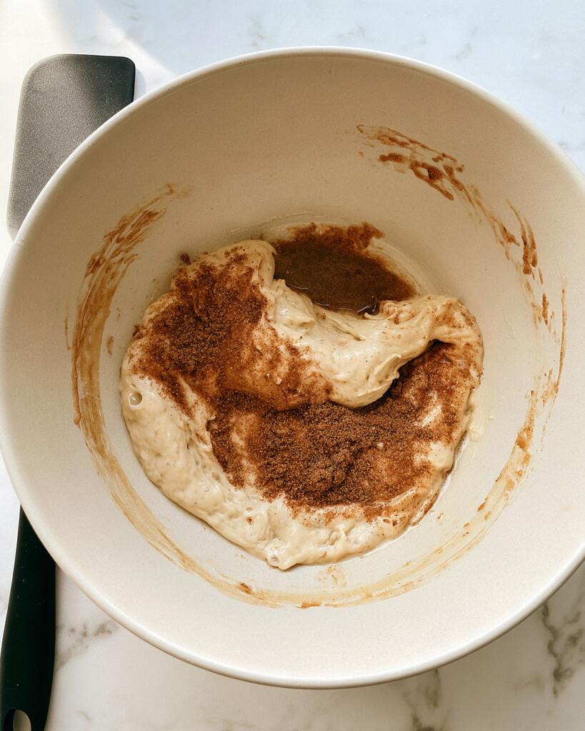 Cinnamon roll focaccia dough inside a white mixing bowl with a visible layer of brown cinnamon sugar on top