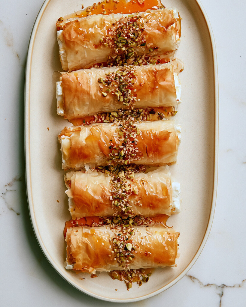 Crispy phyllo feta rolls arranged on a serving plate, topped with a sprinkle of nuts, sesame seeds, and a glossy layer of honey.
