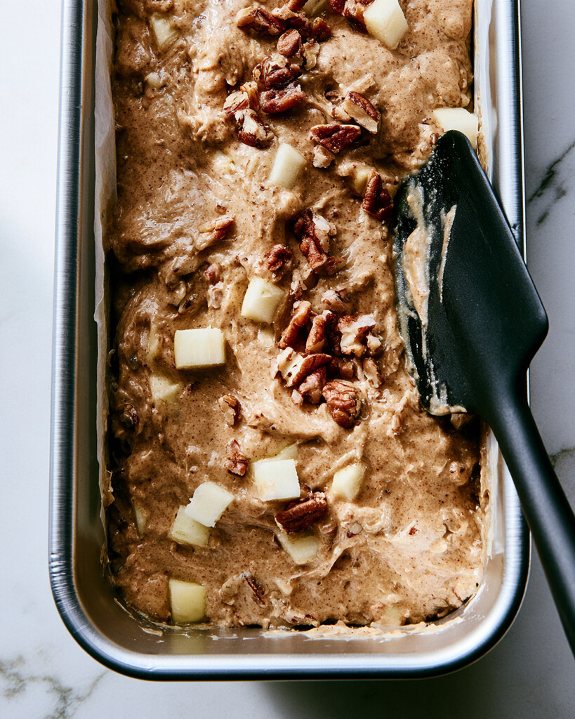 Spiced apple cake batter spread evenly in a parchment-lined metal loaf pan topped with pecans