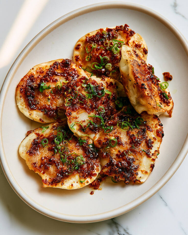 Chili Crisp Smashed Gyoza with Zero Pleating Golden brown open-faced pork dumplings topped with red chili crisp and sliced green onions on a ceramic plate