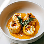 Three brown butter pumpkin ricotta roll-ups on a white plate over orange cream sauce garnished with crispy sage