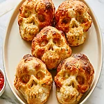Crispy Garlic Butter Pizza Skulls with a Molten Center Five golden brown pizza skulls arranged on a white oval platter next to a bowl of red dipping sauce