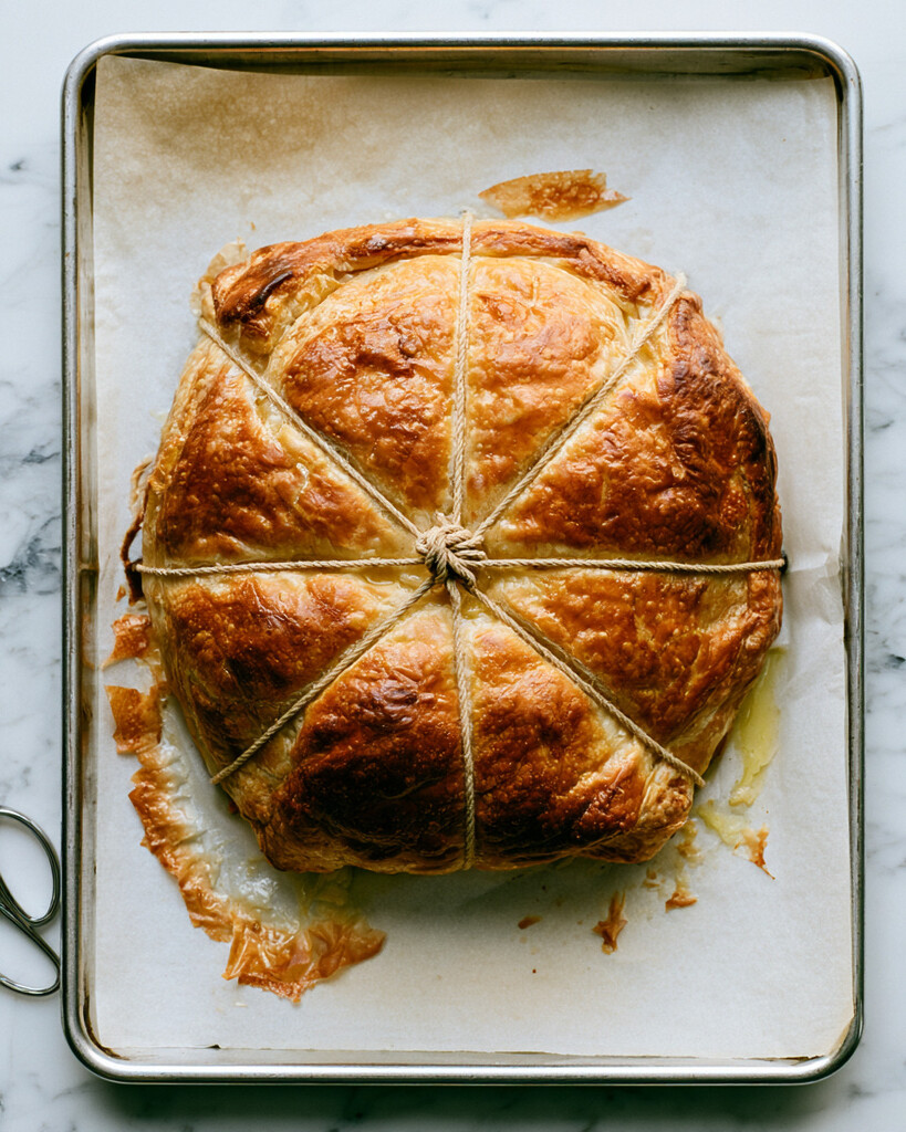 Puffed golden-brown baked brie still tied with kitchen twine resting on a parchment-lined metal baking sheet