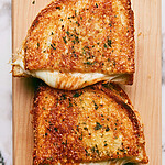 Two garlic bread grilled cheese sandwiches on a wooden board featuring crispy golden parmesan crusts and melted white cheese spilling from the edges