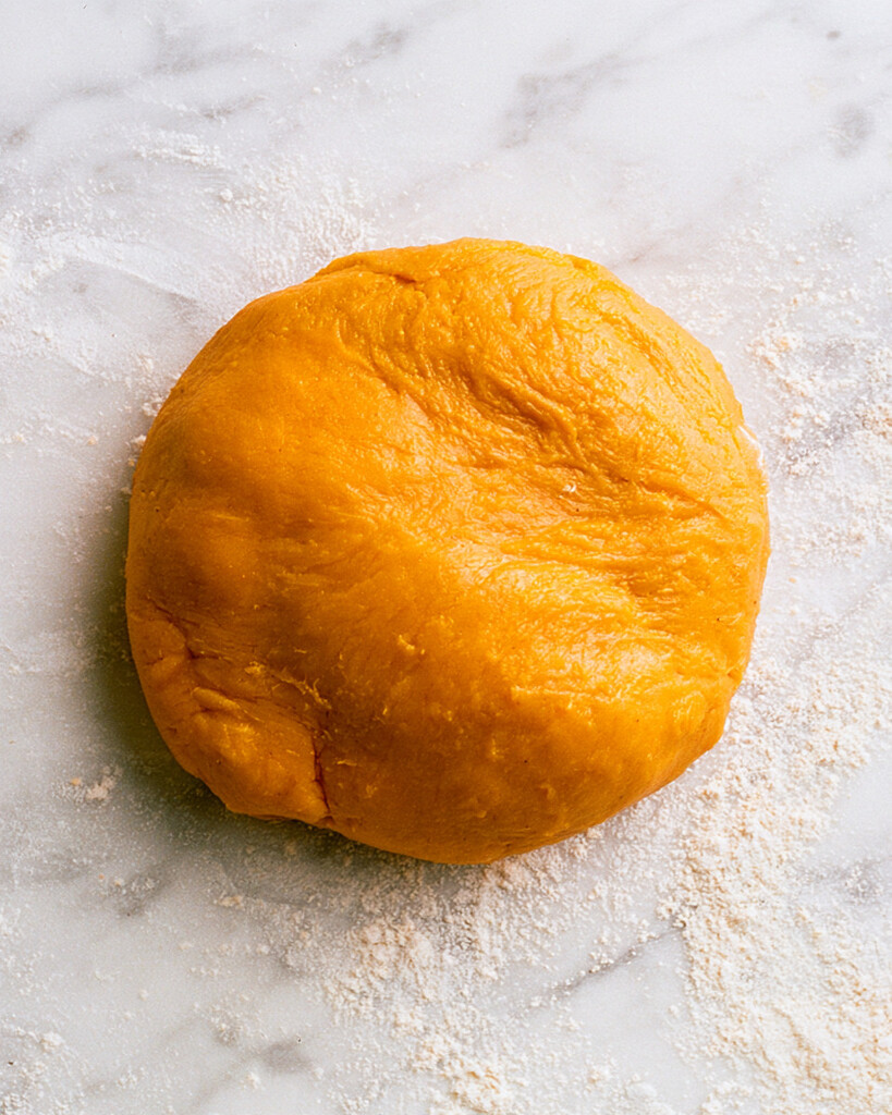 Smooth orange ball of pumpkin gnocchi dough resting on a flour-dusted marble counter